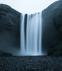 Beautiful Waterfall in Iceland from the front with rough looking water falling off the hill in a dark look with black stones in the foreground and no people in front during night