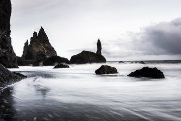 Rocks on a black sand beach in Iceland with reflection in the Sea and a dark sky in misty moody weather with dark colors and a rough sea in landscape format with a storm approaching