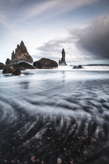 Rocks on a black sand beach in Iceland with reflection in the Sea and a dark sky in misty moody weather with dark colors and a rough sea in landscape format with a storm approaching