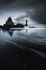 Rocks on a black sand beach in Iceland with reflection in the Sea and a dark sky in misty moody weather with dark colors and a rough sea in landscape format with a storm approaching