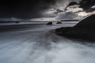 Rocks on a black sand beach in Iceland with reflection in the Sea and a dark sky in misty moody weather with dark colors and a rough sea in landscape format with a storm approaching