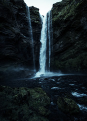 A wild natural fresh sparkling waterfall in a lonely green valley with moos covered rocks and ice cold mountain water flowing down black rocky walls on a rainy moody day in Iceland without tourists