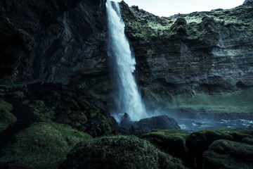 A wild natural fresh sparkling waterfall in a lonely green valley with moos covered rocks and ice cold mountain water flowing down black rocky walls on a rainy moody day in Iceland without tourists