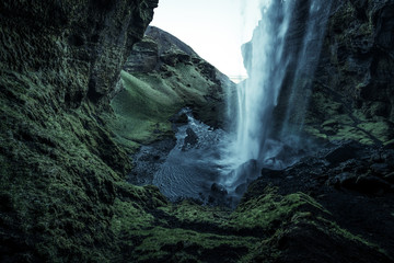 A wild natural fresh sparkling waterfall in a lonely green valley with moos covered rocks and ice cold mountain water flowing down black rocky walls on a rainy moody day in Iceland without tourists
