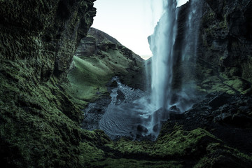 A wild natural fresh sparkling waterfall in a lonely green valley with moos covered rocks and ice cold mountain water flowing down black rocky walls on a rainy moody day in Iceland without tourists
