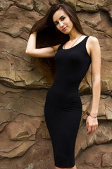 young brunette sexy girl in black dress with long hair is standing with one hand in hair and flirting on stone wall background 