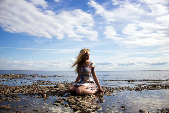 young blonde sexy girl  with long hair in the dress is sitting like nymph on the stones near water to horizont on the blue sky background