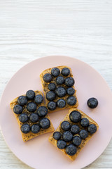 Vegetarian toasts with peanut butter, blueberries and chia seeds on a pink plate over white wooden surface, top view. Healthy eating. From above. Copy space.