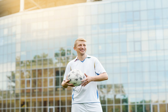 Portrait of a football player against the background of the building in white uniform. Holds a soccer ball. His team won the championship