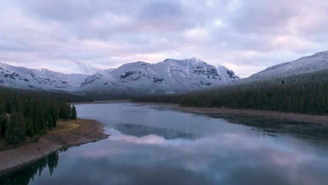 Drone flying over lake toward snow covered mountain at sunset.