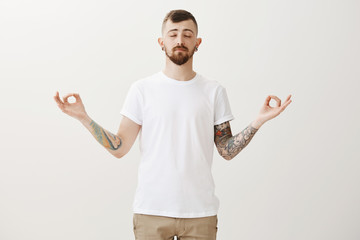 Need to stop and calm down. Portrait of relaxed good-looking urban guy with tattoos and beard, closing eyes and meditating with spread hands and zen gesture, feeling relaxation during yoga lesson