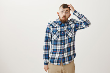 Studio shot of dumb handsome man in plaid shirt and falling glasses, scratching head and folding lips while saying wow, being confused and clueless during math classes, having no idea about topic