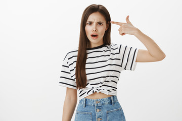 Are you insane or out of your mind. Portrait of outraged shocked european female in striped t-shirt, holding index finger near temple, frowning and arguing, being angry to hear stupid things