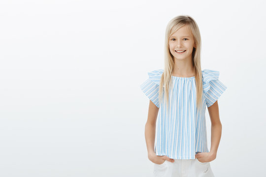 Cute little lady shopping with mom. Studio portrait of pleased happy female child in blue blouse, holding hands on hips and smiling broadly, being amazed, having fun with friends over gray background