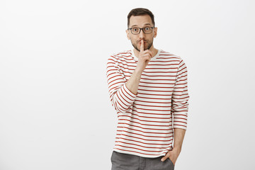 Portrait of creative good-looking adult male designer in striped shirt, asking keep secret, shushing and making shh gesture with index finger over mouth, being intrigued and happy over gray wall