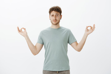 Studio shot of unfocused funny adult man with bristle in earrings, peeking with one eye while standing with spread hands in zen gesture and practicing yoga or meditation, being intense