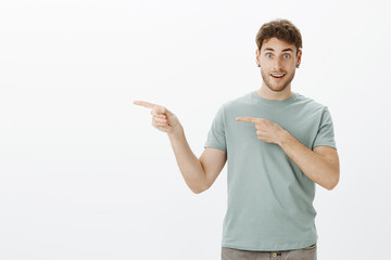 Excited good-looking mature guy in earrings and t-shirt, pointing left with index fingers and smiling broadly, asking if friends want go with him, having desire to visit cool store over gray wall