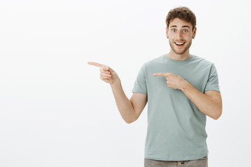 Happy handsome young man with earrings, pointing left with index fingers and smiling joyfully, showing something awesome and interesting, grinning from positive emotions over gray background