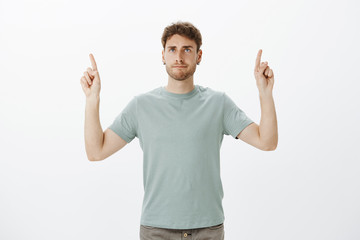 Casual good-looking male coworker in t-shirt, raising index fingers and pointing up while making indifferent annoyed grimace, being fed up with noise colleague and showing direction office upstairs