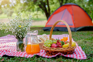 Fototapeta na wymiar Picnic wicker basket with food, bread, fruit and orange juice on a red and white checked cloth in the field with green nature background. Picnic concept.