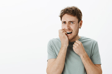 Portrait of stressed anxious european male with fair hair, biting fingernail and frowning, grimacing from fright, being nervous and afraid, standing shocked over gray background