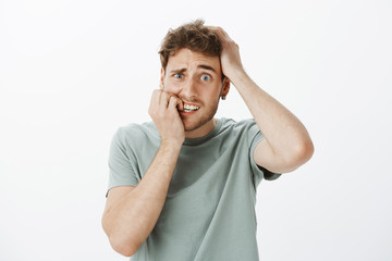 Naklejka premium Guy made mistake feeling afraid what to do. Portrait of shocked nervous Caucasian man with fair hair, holding hand on head and biting fingernail, clenching teeth, being scared over grey wall