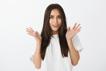 No way, shocking news. Portrait of impressed attractive european woman in white t-shirt, raising palms and gazing at camera with broad positive smile, being fascinated and surprised over gray wall