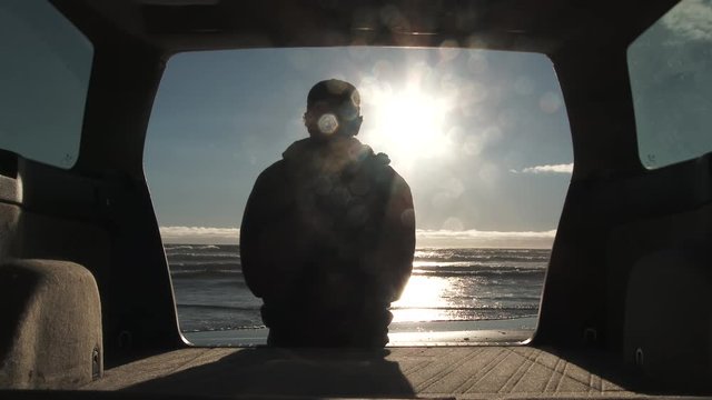 Silhouetted person sitting on tailgate of car watches the waves break on the beach on sunny day in Oregon, then gets up and walks out of frame. Model released for commercial use.
