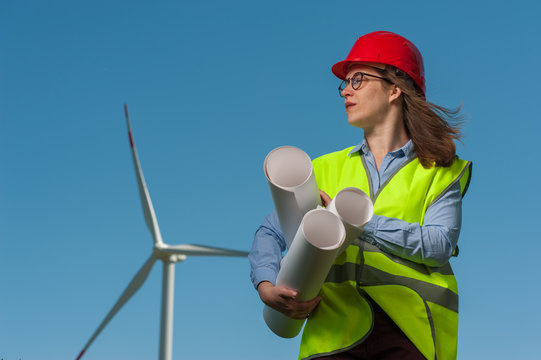Portrait of a serious cute young female engineer in a green waistcoat and red hard hat with geodesic plans in hand against the backdrop of a windmill and blue sky.