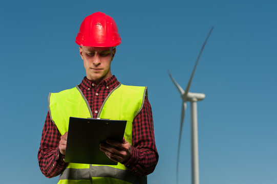 Young handsome male chief engineer in a green waistcoat and red protective helmet signs a design of engineering works against the backdrop of a windmill and a blue sky.