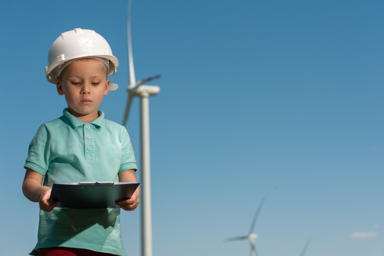 Portrait of a serious ambitious little boy with a helmet with an engineering plan in his hands against the backdrop of a windmill and a blue sky.