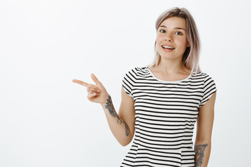 Have you met my friend. Portrait of good-looking friendly female in striped t-shirt with tattooed arms, pointing left with finger gun and smiling joyfully with braces on teeth, posing over grey wall