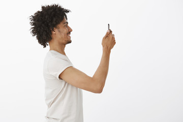 Let me take picture. Profile shot of carefree happy handsome man with bristle and afro haircut, raising hand with smartphone and smiling broadly while photographing girlfriend, standing over gray wall
