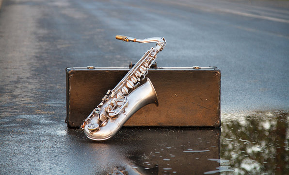 Silver saxophone based on a suitcase standing on an empty, asphalt, wet road