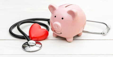 Piggy bank with stethoscope and red heart on white wooden table.