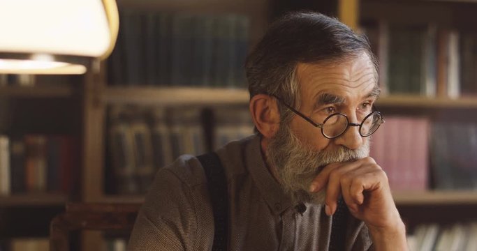 Close up of the old man in glasses and with gray beard being in the library rom while considering about something. Portrait shot.