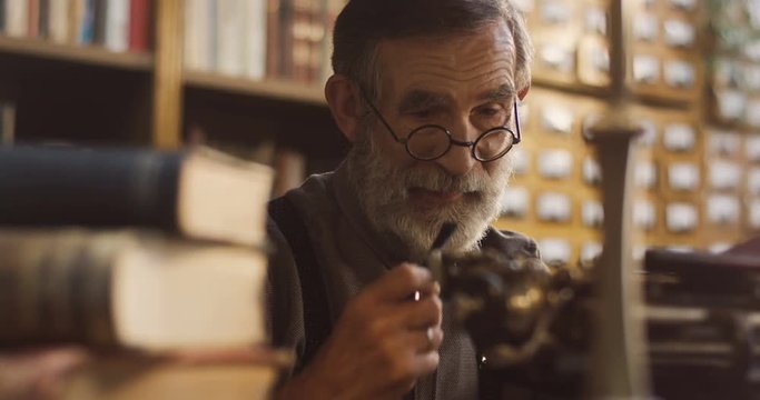 Close up of the senior gray haired man in glasses smoking a tabacco pipe while working on the typewriter in the library room among books.
