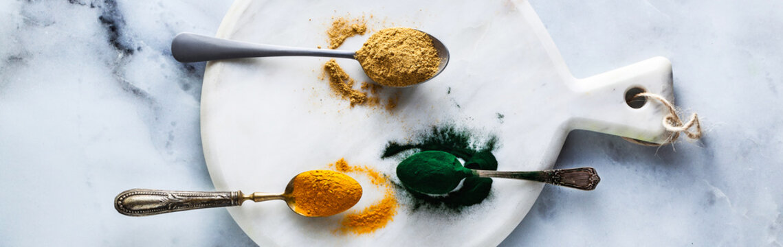 banner of ginger and turmeric powder and hawaiian spirulina in tablespoons on the board and white marble table. healthy supplements, superfoods in cooking
