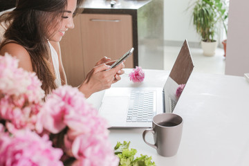 brunette girl typing a message in the phone next to the laptop. enter the password in your phone to pay the bill or buy a plane ticket. girl at the Desk with peonies and pink flowers