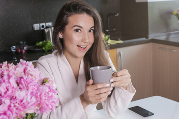 a young girl in a Bathrobe drinking morning tea or coffee from a gray Cup. On the table is a vase with peonies and a phone