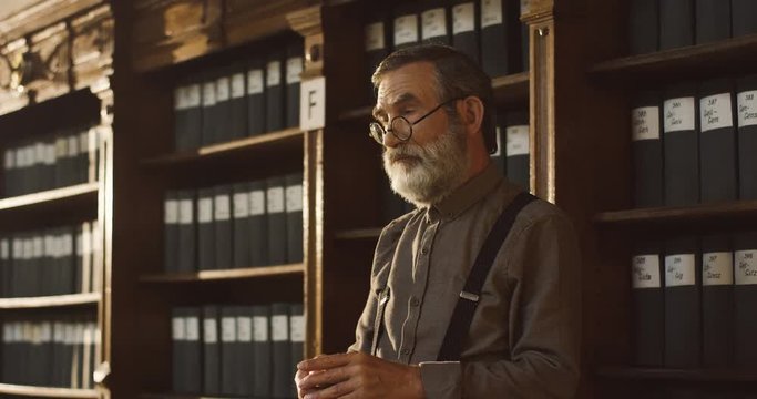Portrait shot of the Caucasian old professor in glasses walking in the library room and checking time on the watch on a chain.