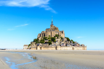 General view of the Mont Saint-Michel tidal island, located in France in Normandy, from the bay at low tide under a summer blue sky with a small stream winding in the sand in the foreground.