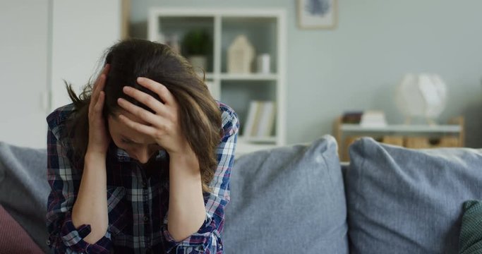 Young woman crying while sitting on the couch at home, then lying on it and pills with water being on the first plan in front of the camera.