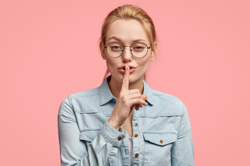 Confident lovely young blonde female shows hush sign, tells secret to someone, touches fore finger on lips, dressed in fashionable denim jacket, has serious expression, isolated over pink background