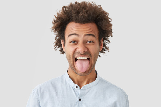 Photo of dark skinned male with Afro hairstyle shows tongue as notices something disgusting, makes grimace, demonstrates stubborn character, dressed in fashionable shirt, isolated on white wall