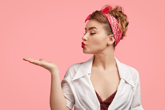 Sideways shot of glamour lady with red lips, clean skin, blows airkiss aside, stretches palm, wears retro outfit, expresses her good attitude and love to someone, poses against pink background