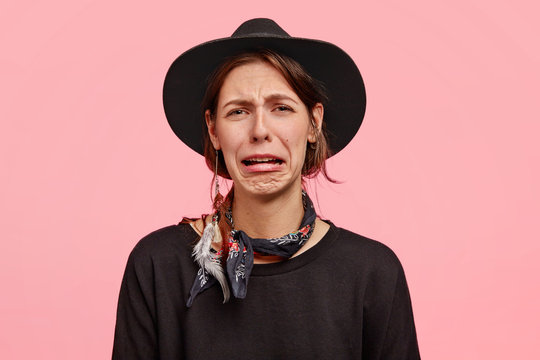 Photo of sorrowful female cries as has grief, purses lips and has discontent facial expression, wears elegant black hat and sweater, poses against pink background. Negative emotions, feelings concept