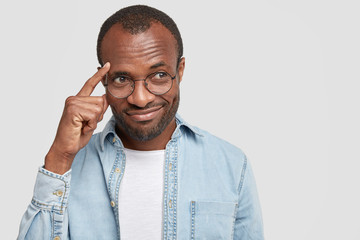 Thoughtful pleased dark skinned middle aged male keeps index finger on temples, tries to concentrate on something, dressed in denim shirt, isolated over white background. Pensive man indoor.