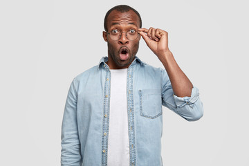 Waist up portrait of stunned unshaven dark skinned male stares through spectacles, dressed in...