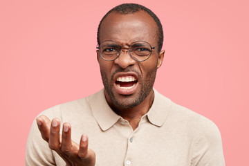 Displeased dark skinned guy frowns face in discontent, feels aversion, shows teeth, stands indoor...